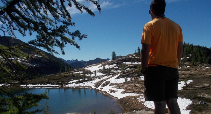 A person faces away from the camera, looking out over a small body of water and a landscape dotted with snow.